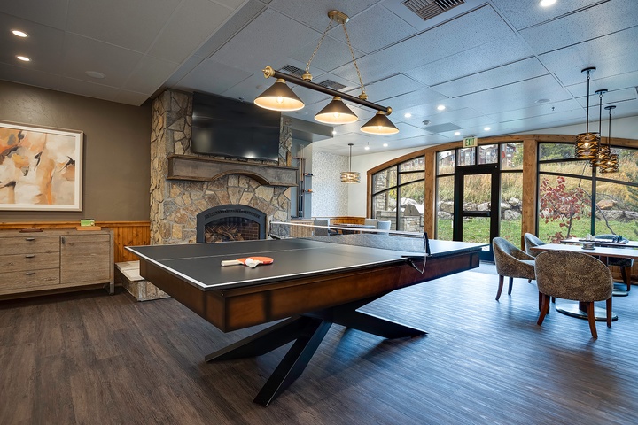 A stylish game room features a ping pong table and stone fireplace, with natural light streaming through arched windows overlooking the scenic grounds.