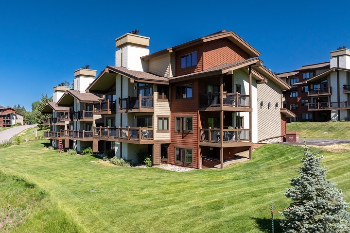 Multi-level alpine-style condominiums with private balconies, nestled in a scenic mountain setting surrounded by manicured lawns and evergreen trees.