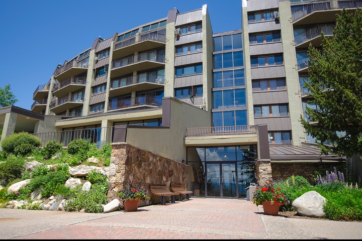 A modern vacation rental property with multiple balconies nestled in a beautifully landscaped garden setting, offering a welcoming entrance amid lush greenery and colorful blooms.