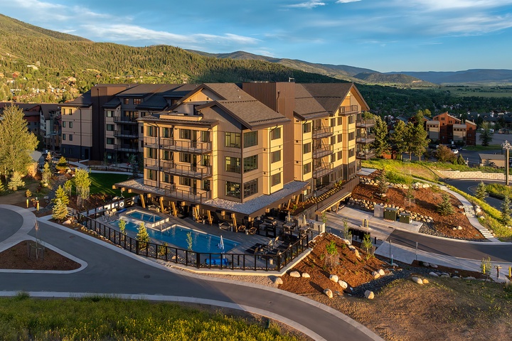 An aerial view captures the property at the base of forested mountains, featuring outdoor pool areas and a multi-story main building nestled within a scenic mountain community. The surrounding hillsides showcase the natural landscape of the area.