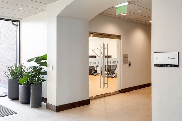 A modern fitness center awaits behind sleek glass doors, with equipment visible and ready for your workout sessions. Fresh greenery adds a welcoming touch to the entrance.