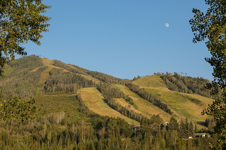 A ski resort nestled in forested mountain slopes offers seasonal recreation under clear skies with the moon visible above.