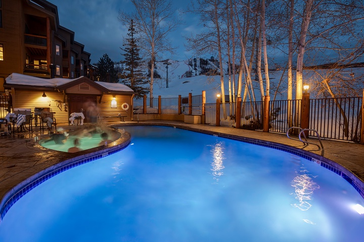 Mountain resort featuring an illuminated outdoor pool and steaming hot tub, nestled among snow-dusted aspen trees with ski slopes visible in the background at twilight.