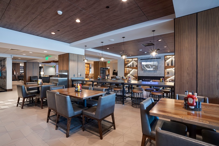 A contemporary communal space invites you to gather and dine with fellow travelers, featuring warm wood tones, modern lighting, and a cozy fireplace visible in the distance.