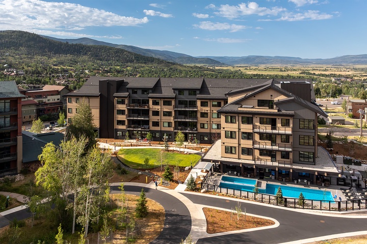 An aerial view of this modern mountain resort property, surrounded by rolling foothills and distant peaks. The well-appointed grounds feature lush lawns, outdoor pools, and scenic valley vistas stretching beyond the property.