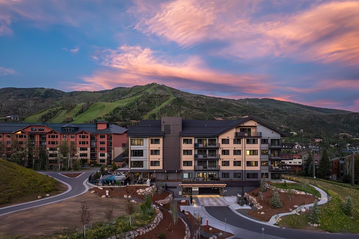 A resort property with modern multi-story buildings and ski slopes nestled at the foot of green mountains beneath a colorful sunset sky.