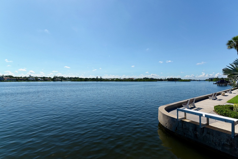 Waterfront seating area overlooking a peaceful lake with distant residential shoreline and blue sky.