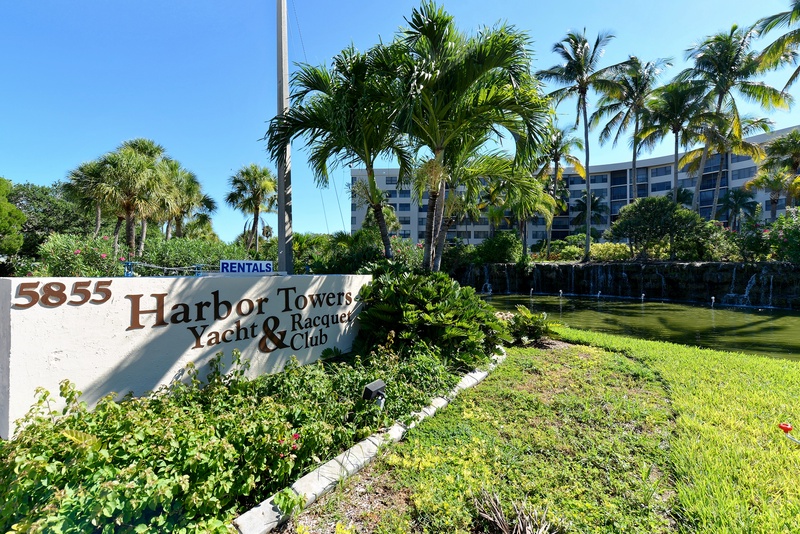 Harbor Towers Yacht & Racquet Club entrance surrounded by lush tropical landscaping and towering palm trees under brilliant blue skies.