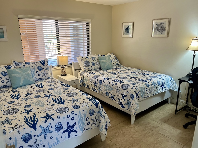 Rest peacefully in your coastal bedroom with twin beds dressed in ocean-themed linens and gentle natural light streaming through blinds.