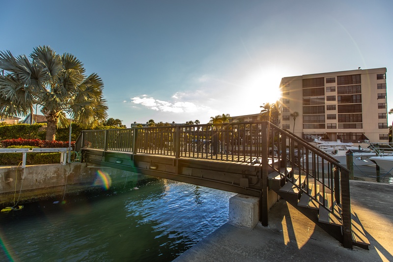 Waterfront bridge connects you to the marina area with tropical palms and nearby property buildings in warm sunlight.