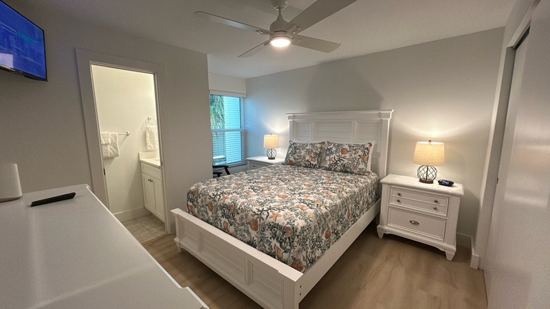 Your peaceful bedroom features a comfortable queen bed with botanical bedding, soft lighting from matching nightstand lamps, and a ceiling fan for your perfect sleep temperature.