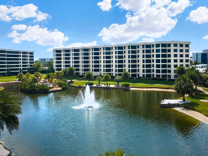 Modern resort buildings overlook a serene lake with decorative fountain, surrounded by tropical landscaping and manicured grounds.