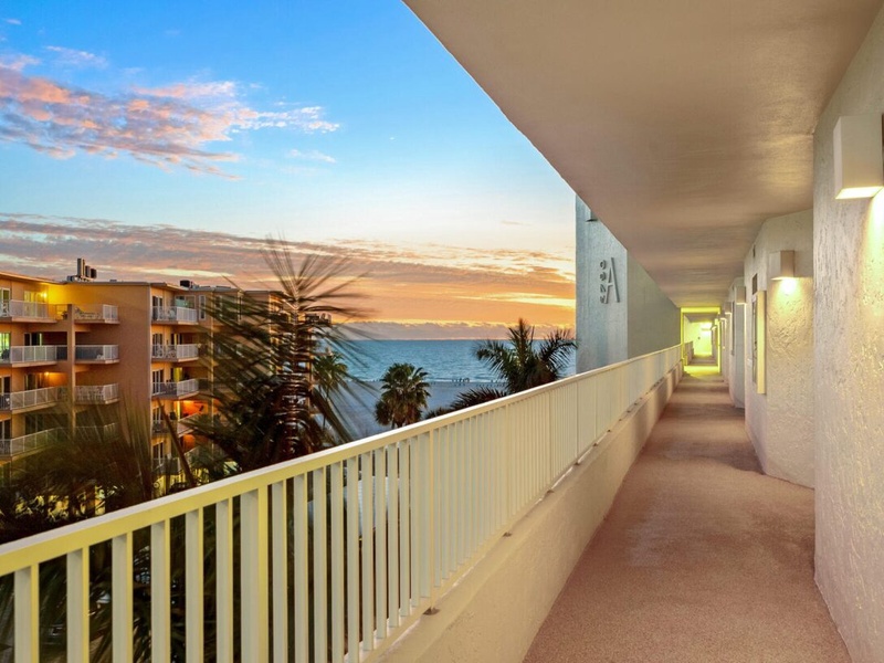 Exterior walkway offers ocean views at sunset with tropical palm trees and neighboring buildings visible.