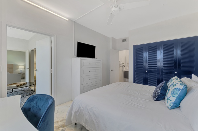 Drift off in your serene bedroom featuring crisp white linens, ocean-blue accents, and modern amenities including TV and ample storage.