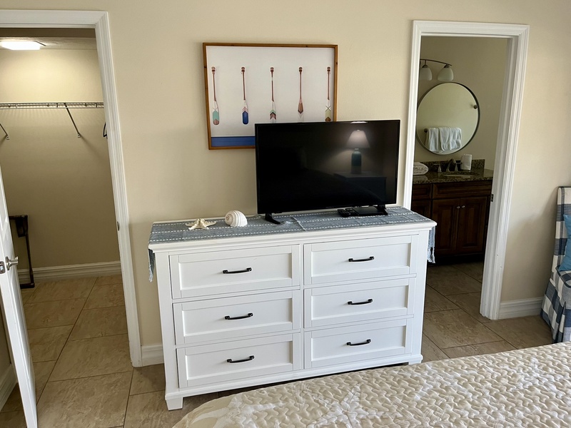 Your spacious bedroom features a white dresser with TV, coastal art, and convenient access to both walk-in closet and ensuite bathroom.