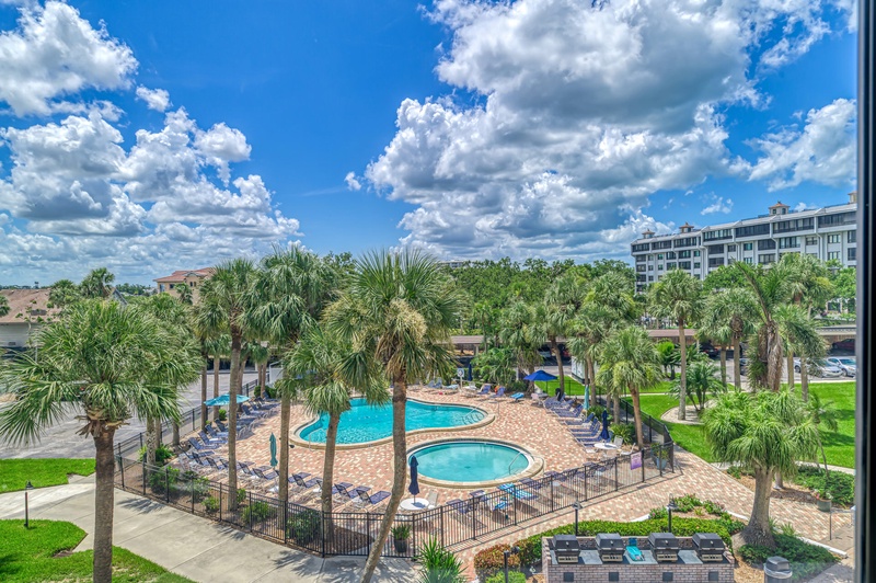 Tropical pool area view featuring palm trees, swimming pools, and modern buildings under a bright blue sky.