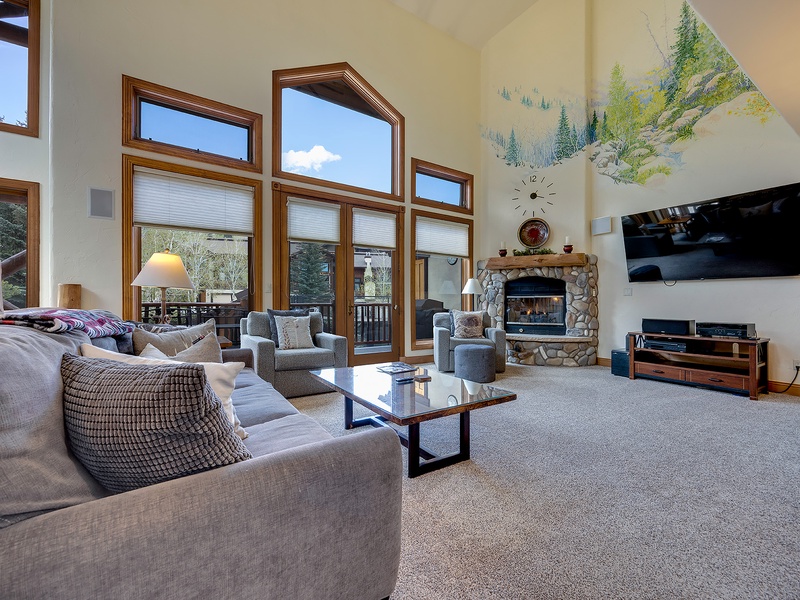 Sink into plush seating where soaring windows frame mountain views, and gather by the stone fireplace as sunlight streams through vaulted ceilings—your alpine retreat awaits.