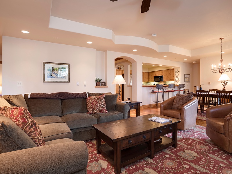 Settle into this spacious living room where plush sofas invite relaxation, warm lighting creates a cozy ambiance, and the open layout connects seamlessly to a full kitchen and elegant dining area—perfect for gathering with loved ones.