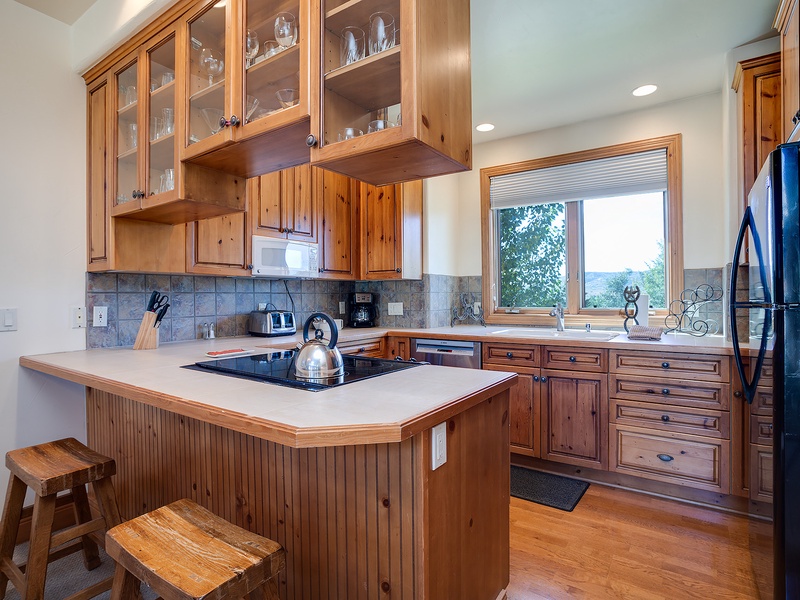 Your fully-equipped kitchen features warm wood cabinetry, modern appliances, and a breakfast bar for casual dining, all framed by stunning mountain vistas through the window.