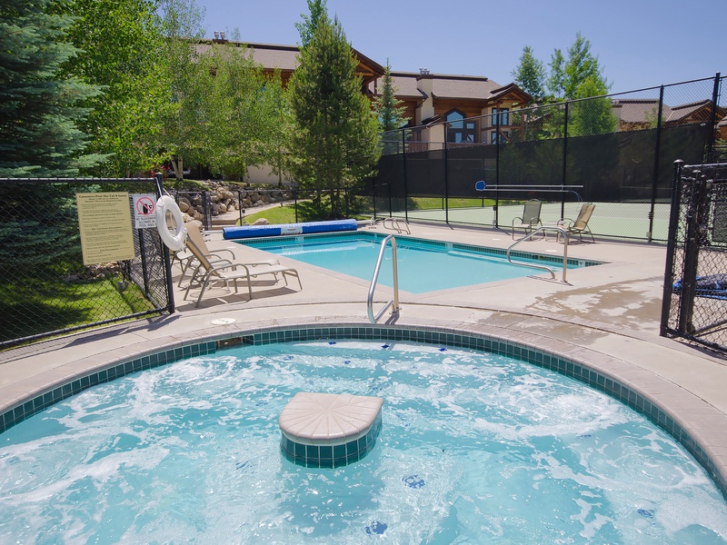 Shared outdoor recreation area featuring heated pool, hot tub, and tennis court surrounded by mature trees and residential buildings in a sunny mountain community setting.