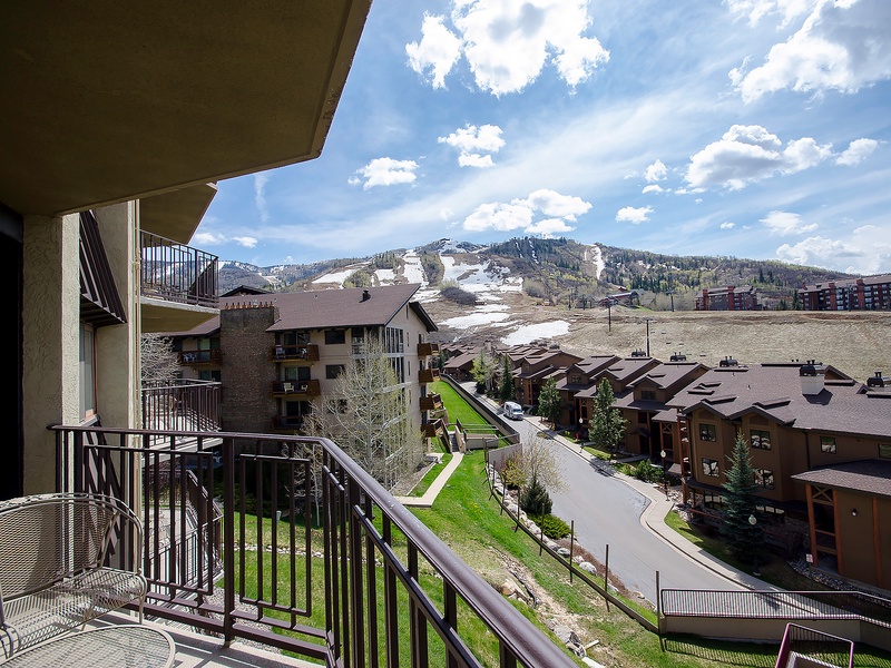 A mountain resort community with ski slopes transitioning from winter, surrounded by residential buildings and natural landscape under partly cloudy skies.