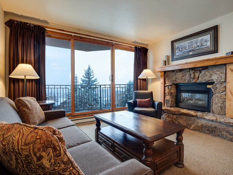 Settle into your mountain retreat with a stone fireplace and floor-to-ceiling windows framing stunning alpine views. Cozy up on plush sofas as natural light fills this welcoming living room.