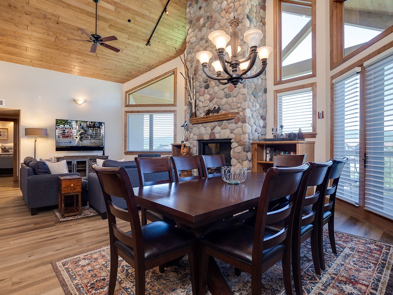 Gather around this handsome dining table beneath soaring wood ceilings and a river-stone fireplace. Natural light pours through floor-to-ceiling windows, framing views of the surrounding landscape while you share meals and make memories with loved ones.