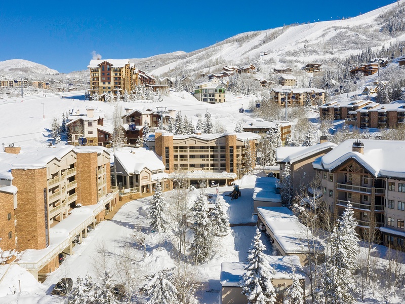 A picturesque alpine village nestled in snow-covered mountains, offering ski-in/ski-out access and a charming mountain retreat. The property sits among scenic slopes and evergreen trees dusted with fresh powder under clear blue skies.