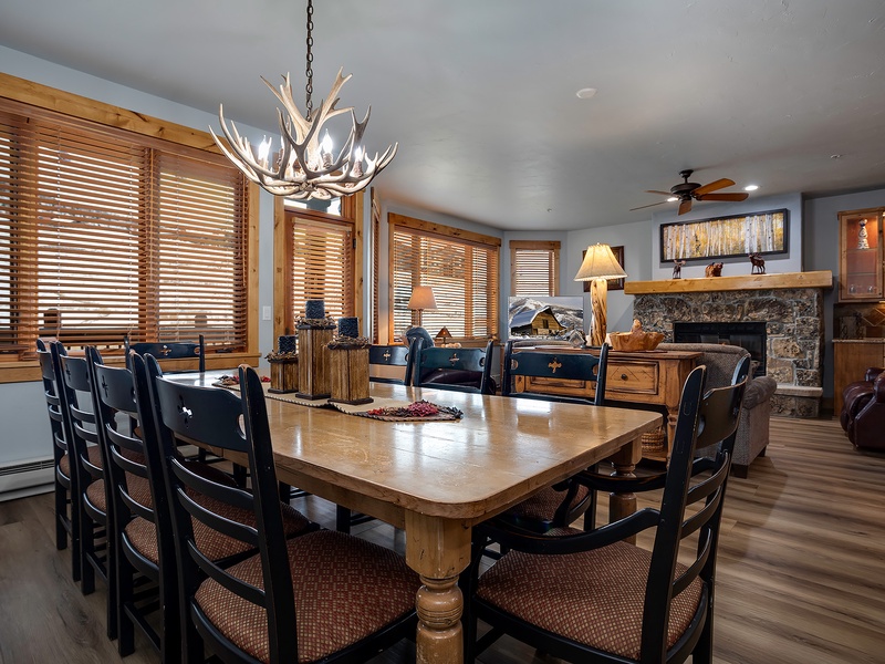Gather around the expansive dining table under a striking antler chandelier, with mountain-inspired décor and a stone fireplace creating a warm, inviting atmosphere for your group meals.