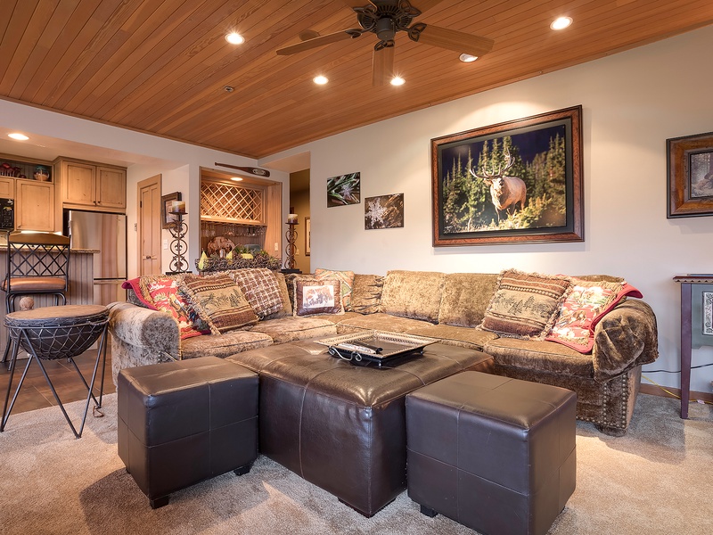 Unwind on the plush sectional beneath warm wood ceilings, where rustic charm meets comfort. Your cozy retreat awaits with inviting seating, nature-inspired art, and an open layout connecting living and dining spaces.