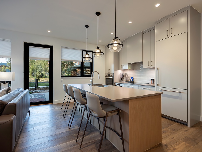 Your sleek modern kitchen invites you to gather around the spacious island, where pendant lighting illuminates a bright space designed for cooking and connection. Natural light streams through glass doors, opening your culinary hub to the outdoors.