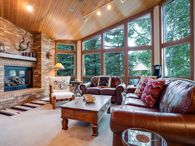 Curl up by the fireplace in this inviting living space, where soaring windows frame lush forest views and leather seating invites you to unwind in natural warmth and comfort.