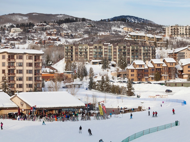 A vibrant winter resort area with ski slopes, lodges, and alpine village buildings nestled among snow-covered mountains and evergreen trees.