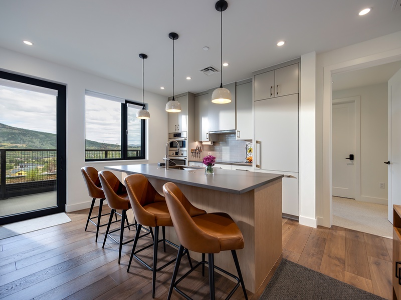 Open-concept kitchen with sleek pendant lights above a spacious island, leather bar stools, and floor-to-ceiling windows framing stunning mountain views.