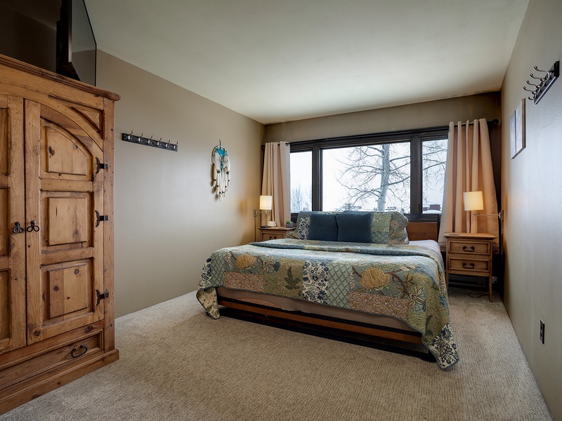 Unwind in your peaceful bedroom with rustic wood accents, plush quilted bedding, and a large window framing serene winter views—your perfect mountain retreat.