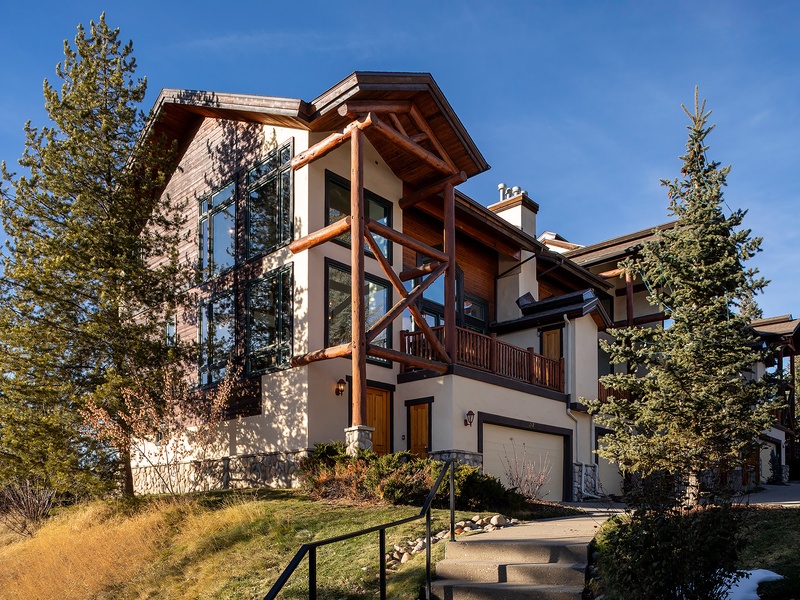 This alpine-inspired mountain retreat features a striking multi-level design with wood and stone elements, nestled among tall evergreens under clear blue skies.