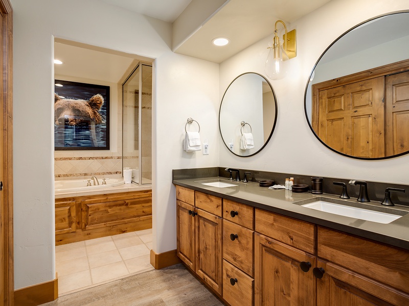Unwind in your private spa-like bathroom with double vanities, elegant round mirrors, and a luxurious soaking tub framed by warm wood cabinetry and charming mountain-inspired artwork.