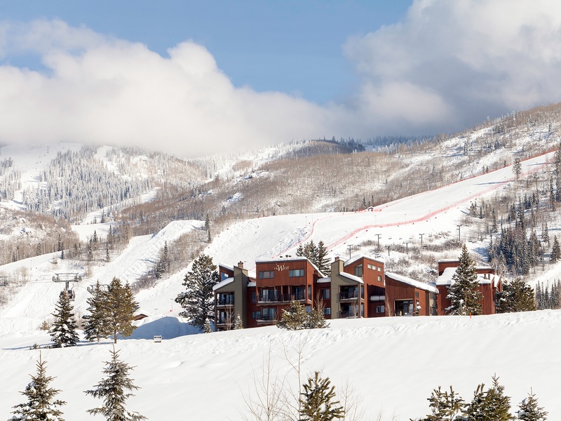 Modern alpine lodging set against a dramatic winter backdrop with ski slopes and snow-covered mountains. The property sits at the base of the resort area surrounded by evergreen trees.