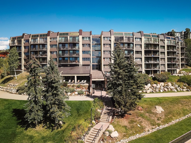 Modern multi-story property nestled in landscaped grounds, surrounded by mature evergreens and natural stone features under a clear blue sky.