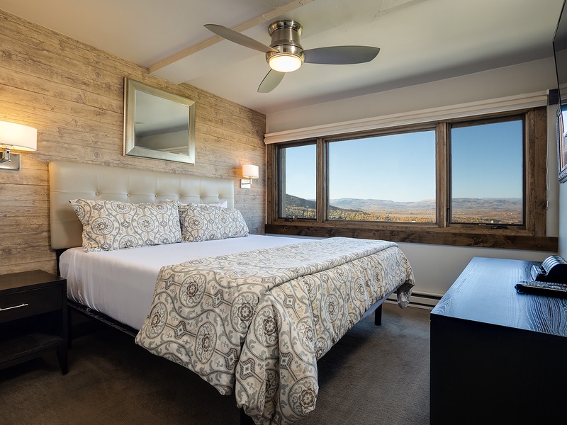 Your serene bedroom features a plush bed with elegant linens, warm wood accents, and stunning panoramic mountain views through expansive windows—perfect for restful mornings.