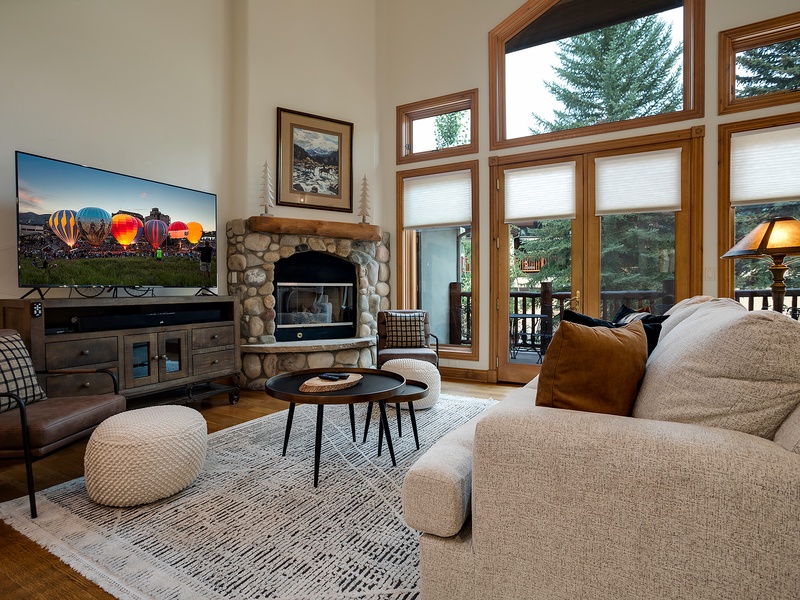 Picture yourself unwinding here—plush seating frames a stone fireplace, while tall windows reveal lush evergreens and a private deck. A cozy space designed for your mountain retreat.