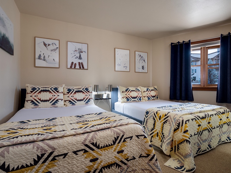Unwind in your cozy mountain retreat with two comfortable beds dressed in geometric patterns, framed alpine art above, and blackout curtains for restful mornings after adventure-filled days.