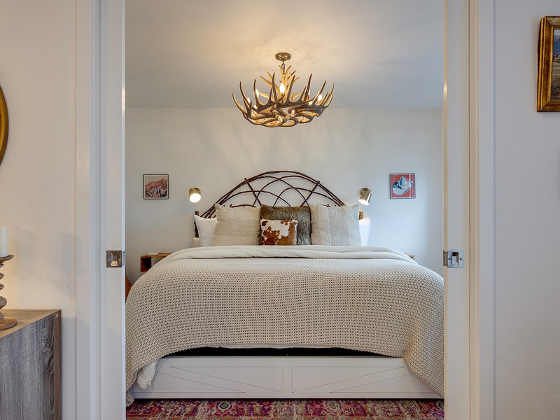 Your bedroom awaits with a plush bed, rustic antler chandelier, and cozy textures. Original artwork and thoughtful touches create a warm, inviting retreat for restful nights.