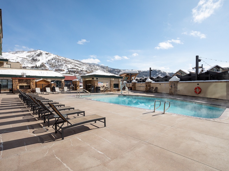 An outdoor pool nestled at the base of snow-covered peaks, with ample loungers and stone accents perfect for après-ski relaxation under clear mountain skies.