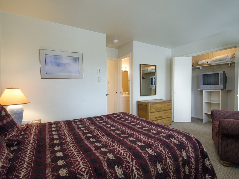 Your cozy bedroom features a plush bed with warm patterned linens, a comfortable armchair, soft lighting, and your own TV. The ensuite bathroom and closet storage keep everything you need close at hand.