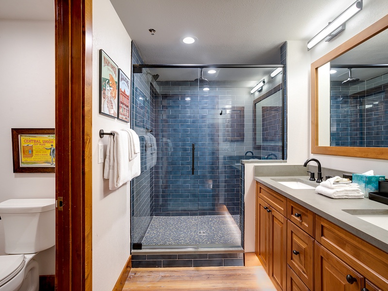 Step into your spa-inspired bathroom featuring a stunning walk-in shower with sleek blue subway tiles, rainfall showerhead, and dual vanity with warm wood cabinetry—perfect for unwinding after your adventures.