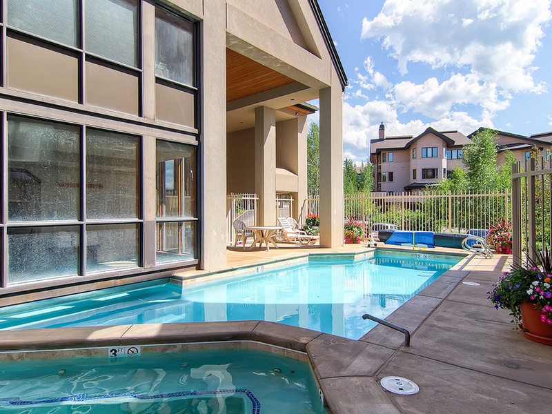 Modern indoor-outdoor pool area featuring crystal-clear water, hot tub, and bright poolside patio with mountain views, all under a dramatic cloud-filled sky in a peaceful residential setting.