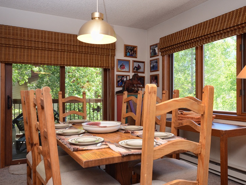 Gather around this warm, inviting dining table where your group can share stories and meals together, surrounded by natural light streaming through large windows that frame lush green views.