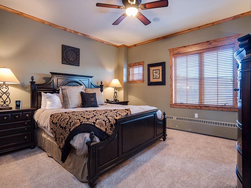 Your cozy bedroom features a plush queen bed with elegant linens, warm bedside lamps, and a ceiling fan for your comfort. Large windows invite natural light into your serene mountain retreat.