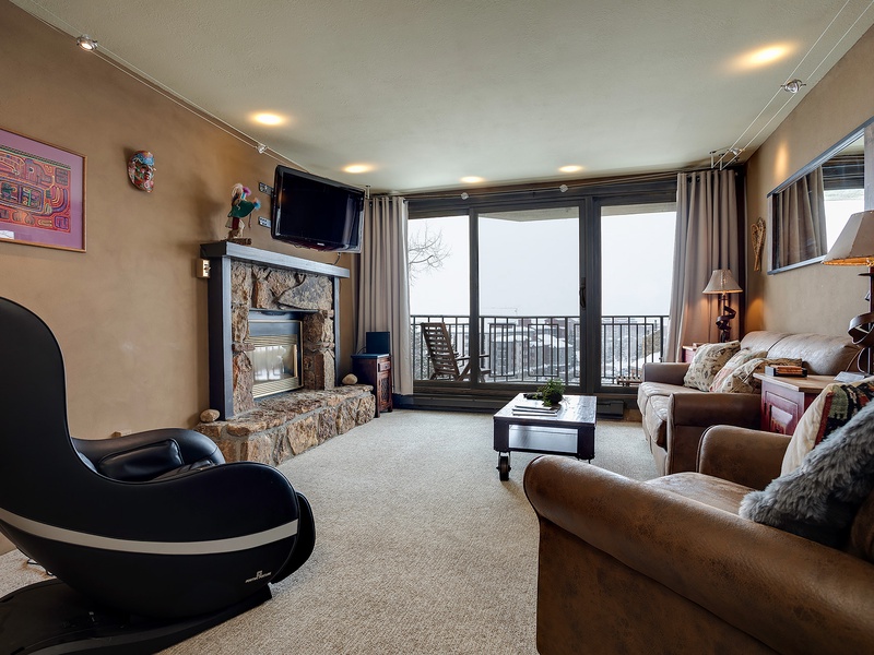 Your inviting living space features plush sofas, a cozy stone fireplace, and a private balcony where you can unwind while enjoying scenic mountain views through floor-to-ceiling windows.